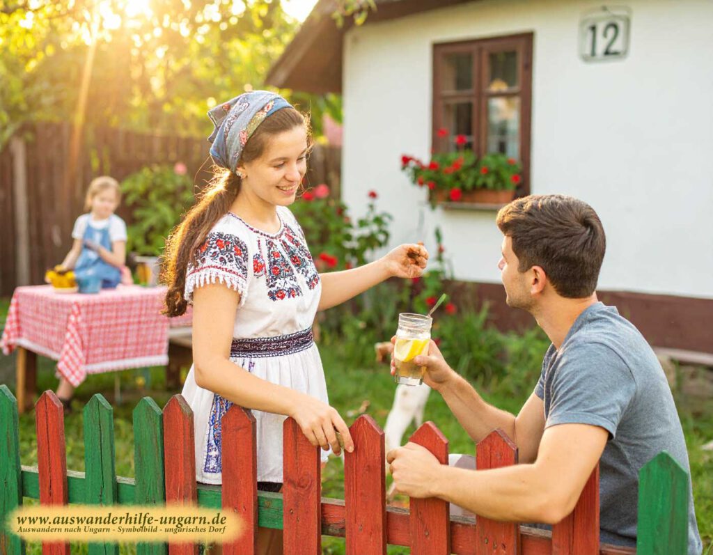 Junge Frau mit bestickter Bluse reicht einem Mann am rot-grünen Holzzaun ein Glas Limonade; im Hintergrund Kind an einem rotkarierten Tisch vor Dorfhaus Nr. 12 mit Geranien – warmes Abendlicht in Ungarn.