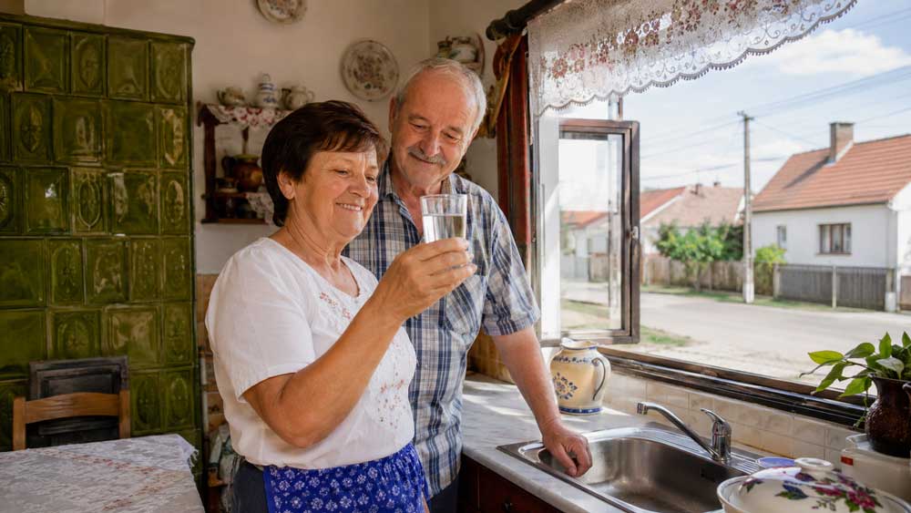 Älteres Paar betrachtet in einer ungarischen Küche ein Glas Wasser am Spülbecken mit Blick auf Dorfstraße und Stromleitungen passend zu Auswandern nach Ungarn und Energieversorgung
