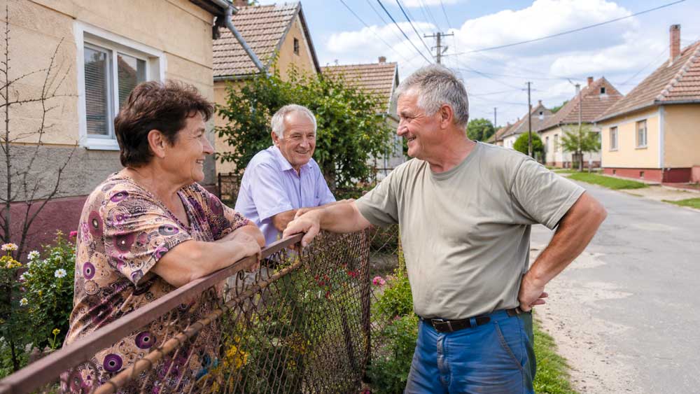 Drei Erwachsene unterhalten sich freundlich am Gartenzaun vor ungarischen Dorfhäusern mit Stromleitungen und ruhiger Straße