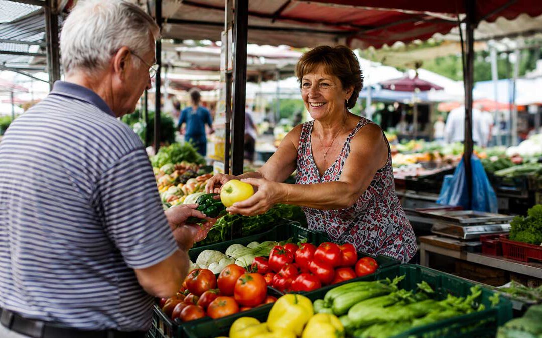 Älterer Mann kauft auf ungarisch frisches Gemüse an einem Marktstand; eine Verkäuferin reicht ihm Paprika und Gurken zwischen Kisten mit Tomaten, Paprika und anderem Gemüse.